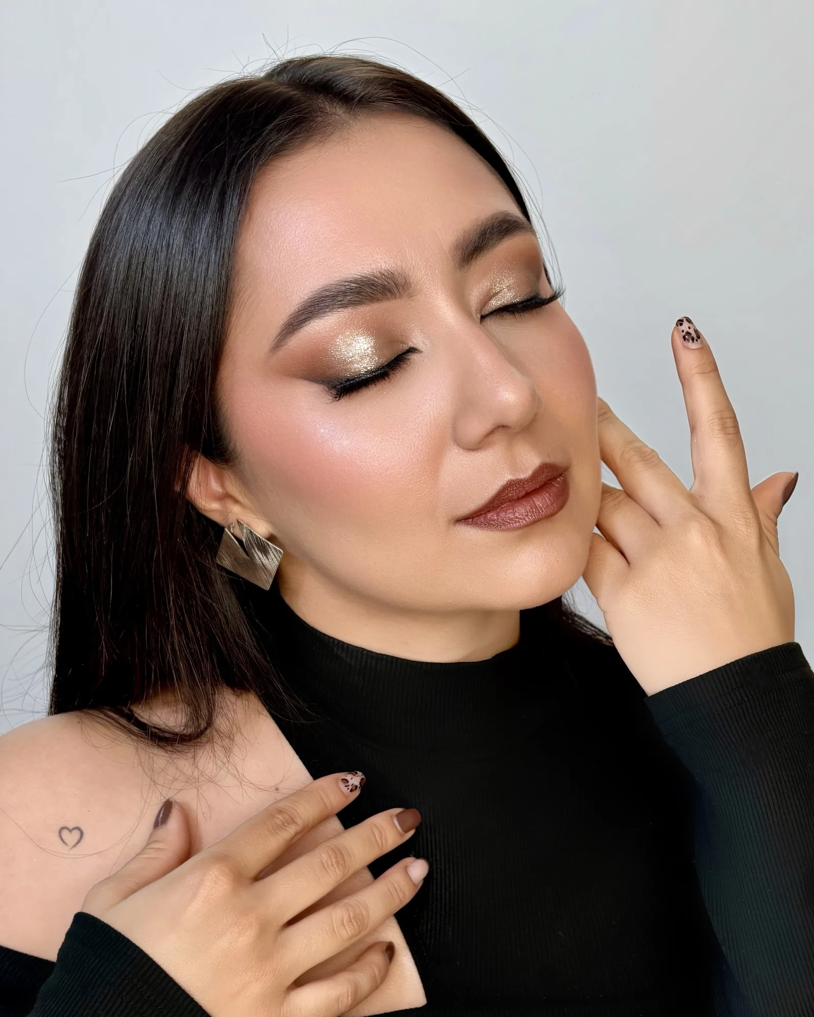 Maquillaje social con sombra dorada en los párpados, piel luminosa y labial vino, con la mano junto al rostro