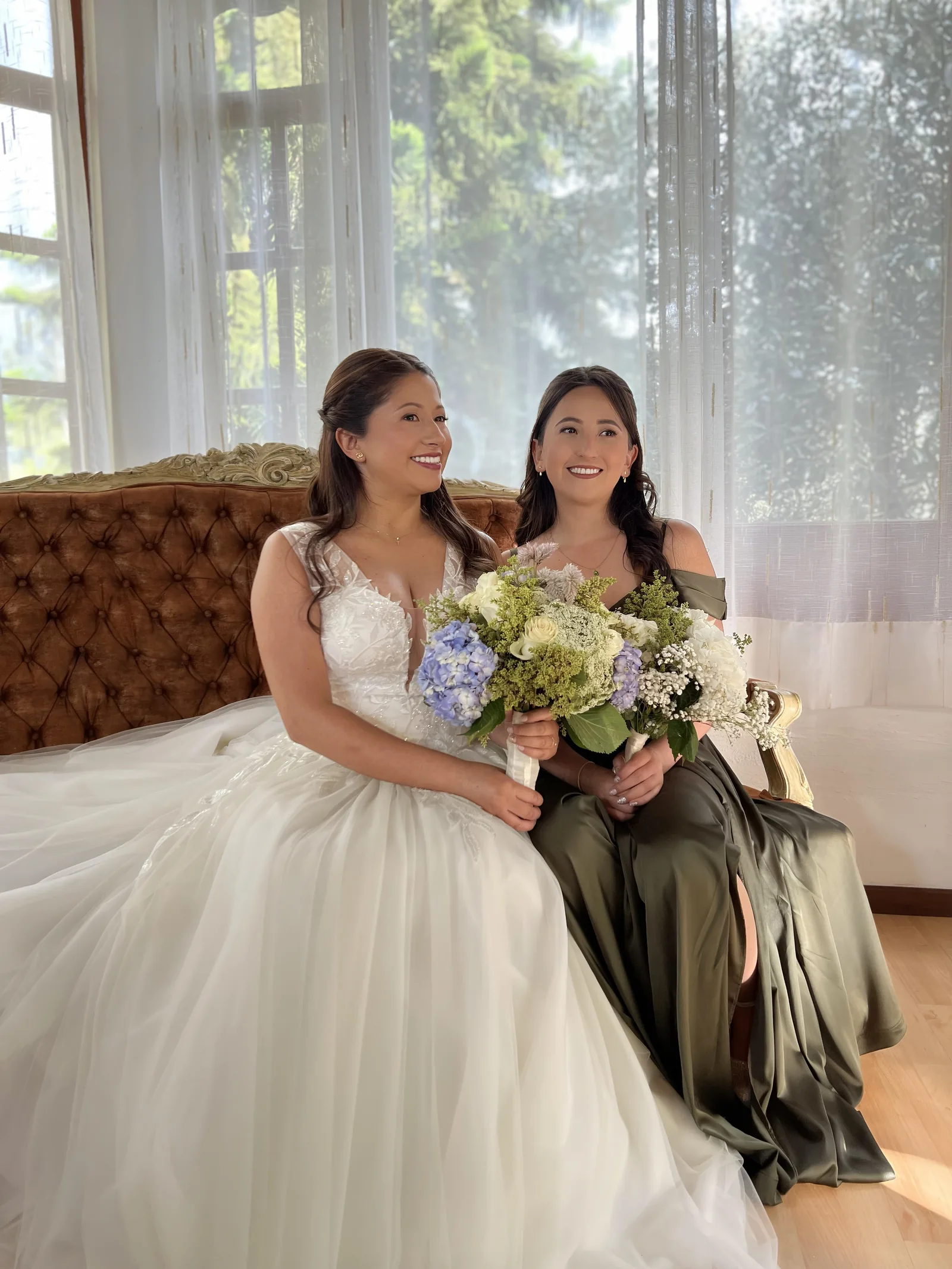 The bride and a companion pose with a bouquet by a window.