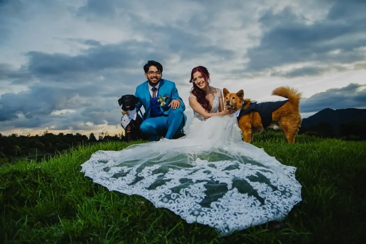 Andre con maquillaje de novia y ondas rojizas junto al novio y dos perros, con velo de encaje en el pasto