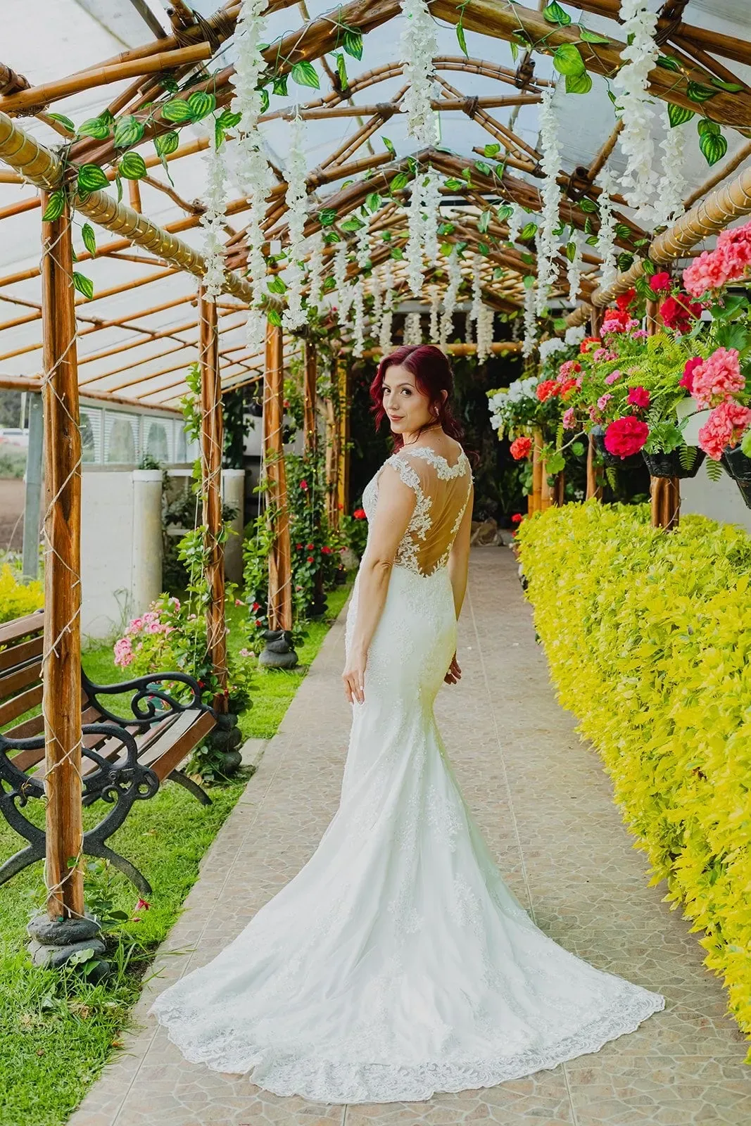 Andre mira atrás con maquillaje de novia y ondas rojizas, en vestido sirena bajo una pérgola florida