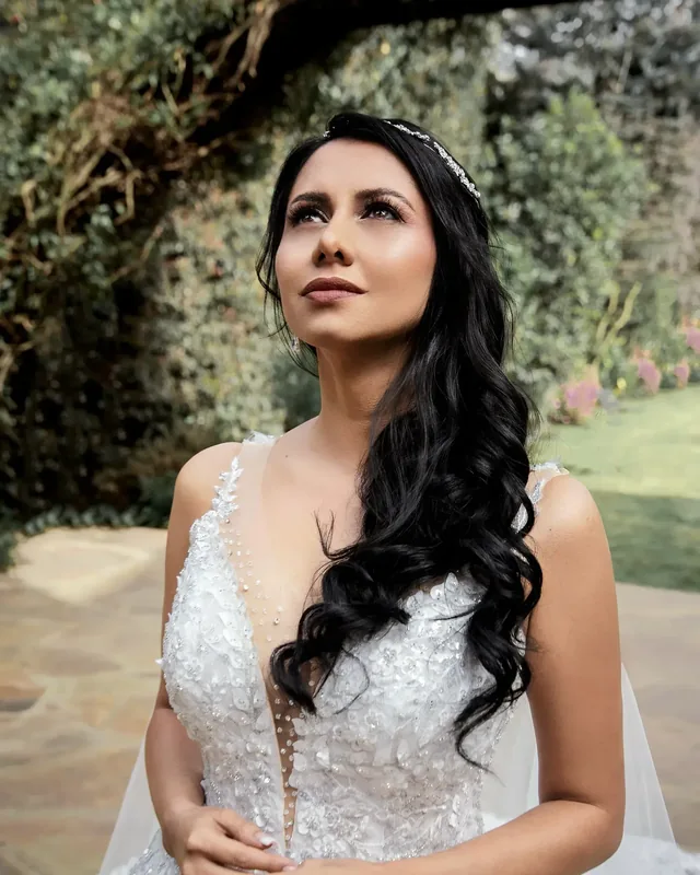 Caro mira hacia arriba en un jardín, con maquillaje de novia de piel luminosa, ondas largas y tiara fina