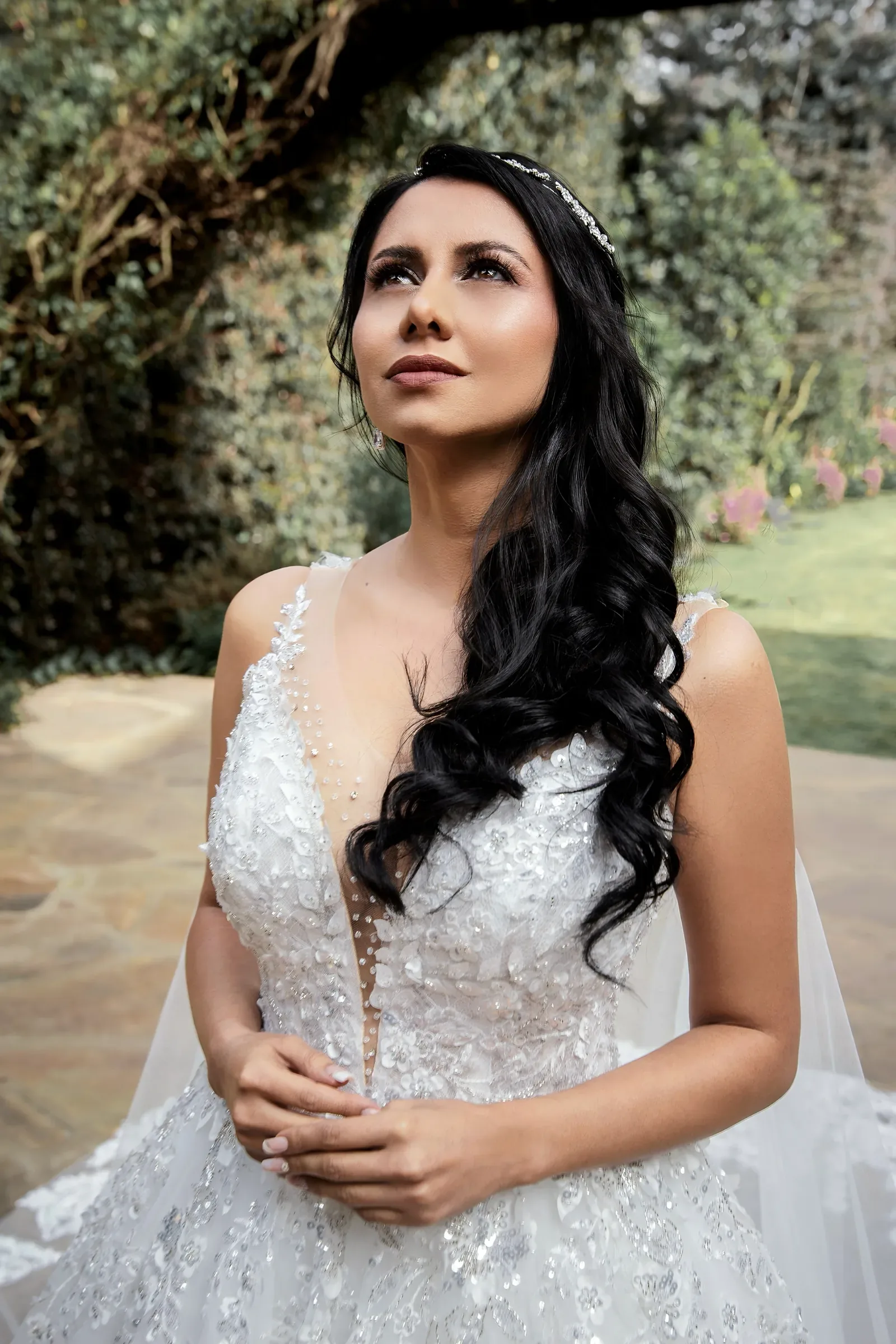 Caro mira hacia arriba en un jardín, con maquillaje de novia de piel luminosa, ondas largas y tiara fina