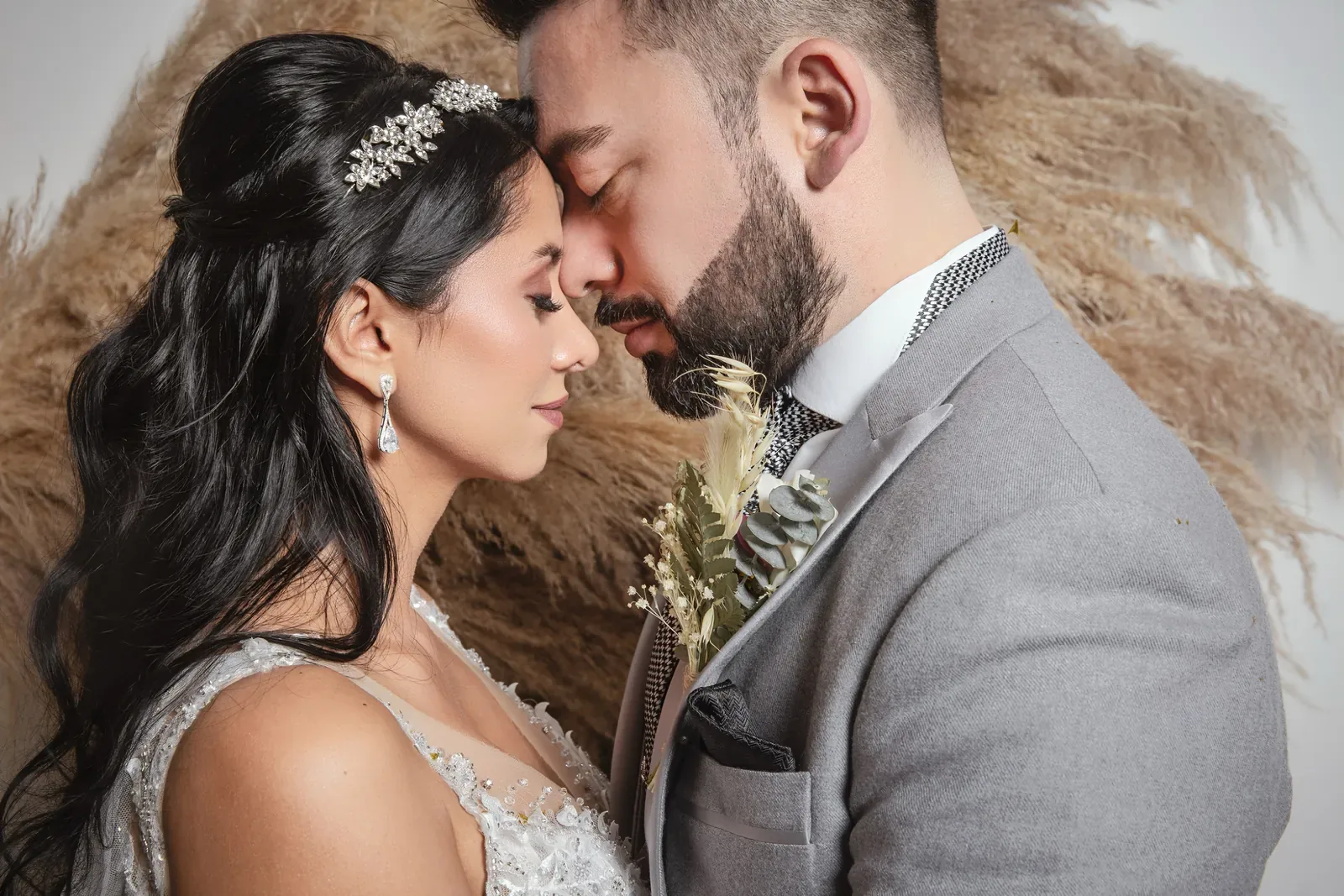 Caro, con maquillaje de novia luminoso y tiara, junta su frente con la del novio frente a plumas de pampa