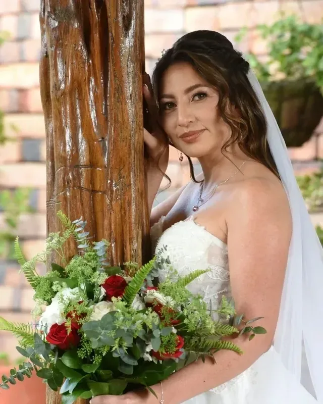 Erika con maquillaje de novia de piel luminosa, recostada en una columna de madera con ramo de rosas rojas