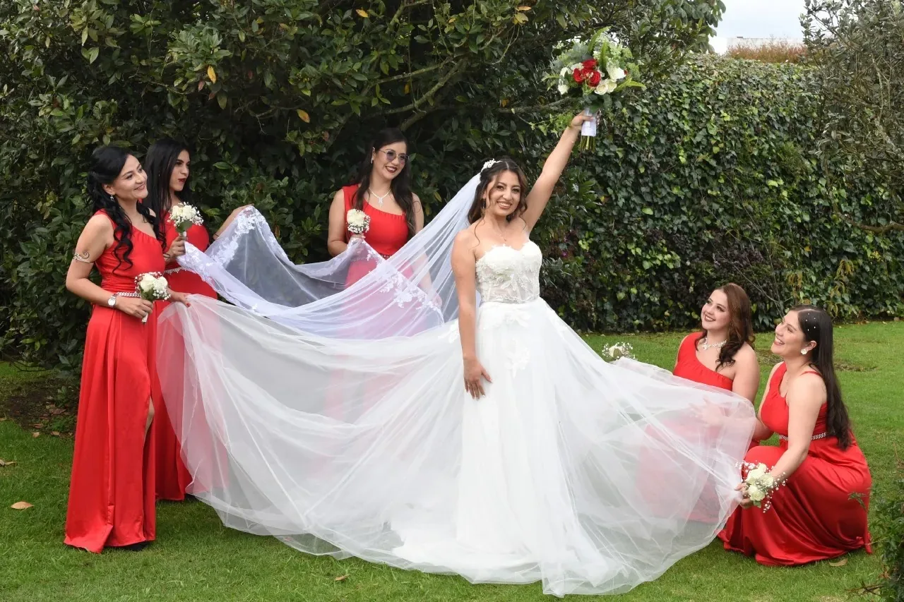 Erika con maquillaje de novia alza su ramo mientras cinco damas en vestidos rojos extienden su velo