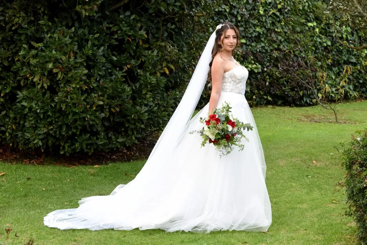 Erika con maquillaje de novia de pie en un jardín, con velo largo, vestido de tul con cola y ramo rojo