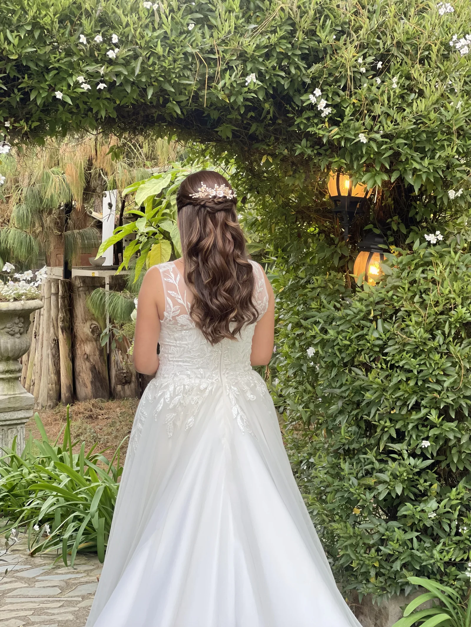 Bride seen from behind in an embroidered white gown, with wavy hair and a floral hairpiece in a garden.