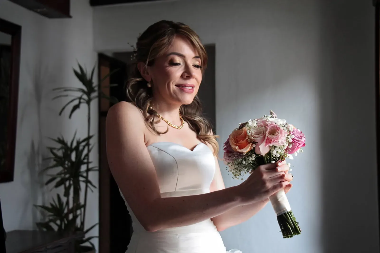 Ginita sonríe mirando su ramo de rosas, con maquillaje de novia dorado y vestido strapless de escote corazón