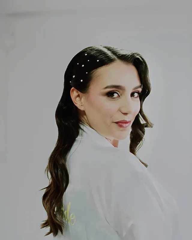 Katha mira sobre el hombro con maquillaje de novia de ojos cálidos, ondas largas y perlas en el cabello