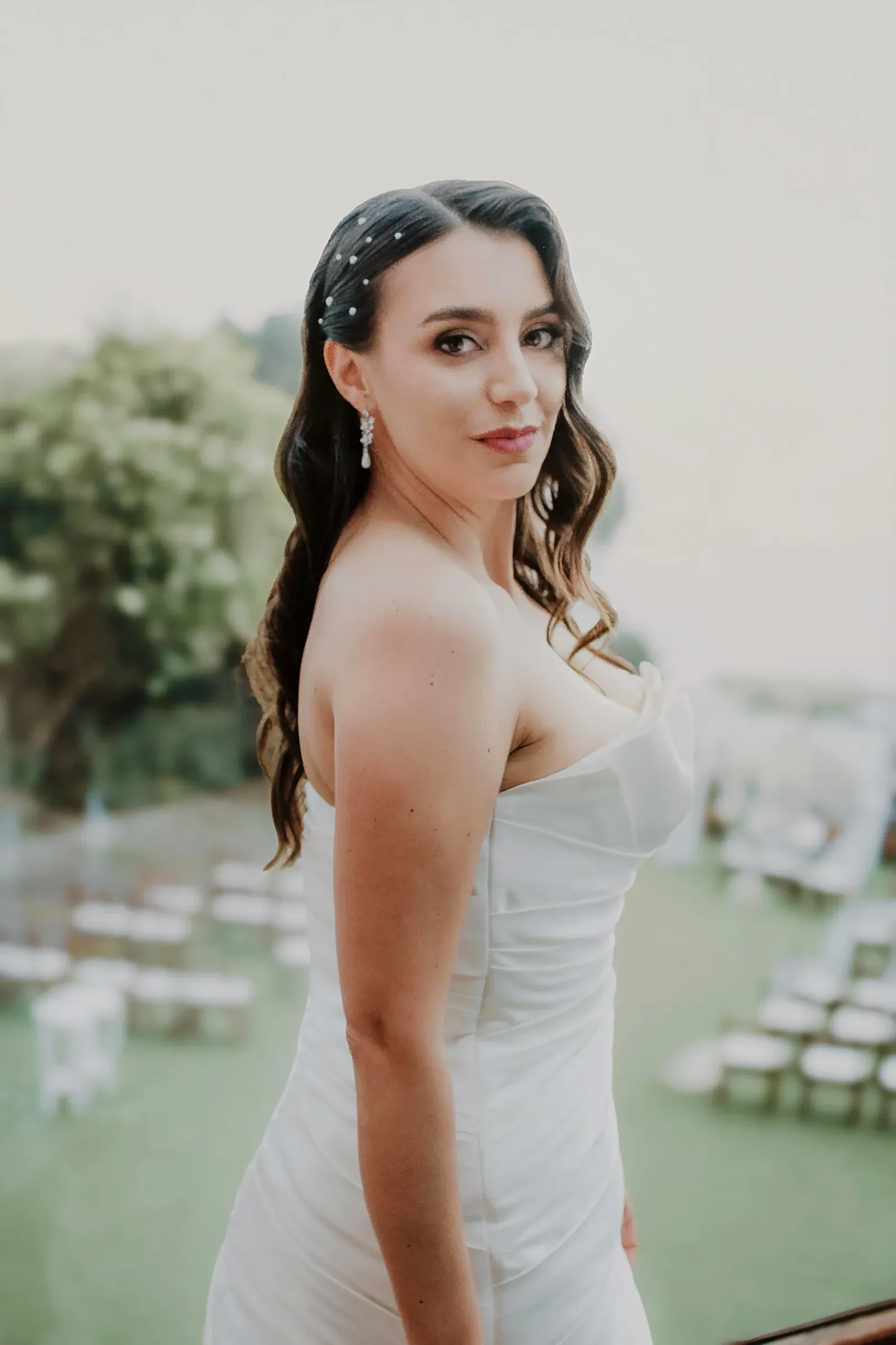 Katha con maquillaje de novia, ondas largas con perlas y vestido strapless en una terraza sobre un jardín