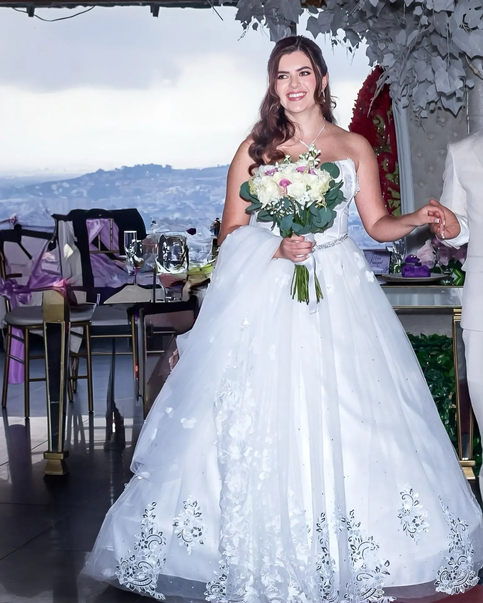 Kelly sonríe con maquillaje de novia natural, cabello suelto en ondas y ramo de rosas blancas