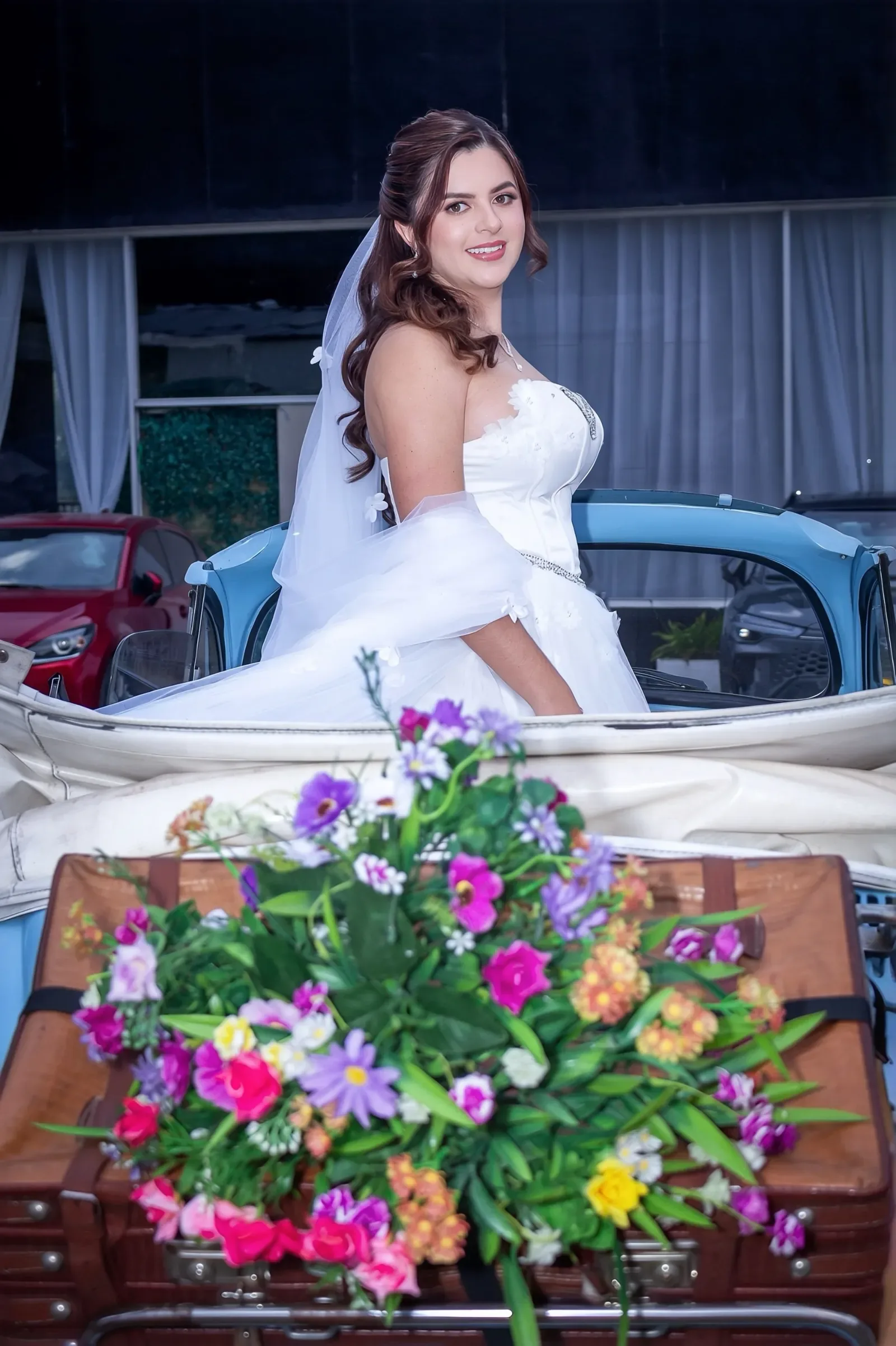 Kelly con maquillaje de novia luminoso, semirrecogido en ondas y velo, de pie en un auto clásico azul