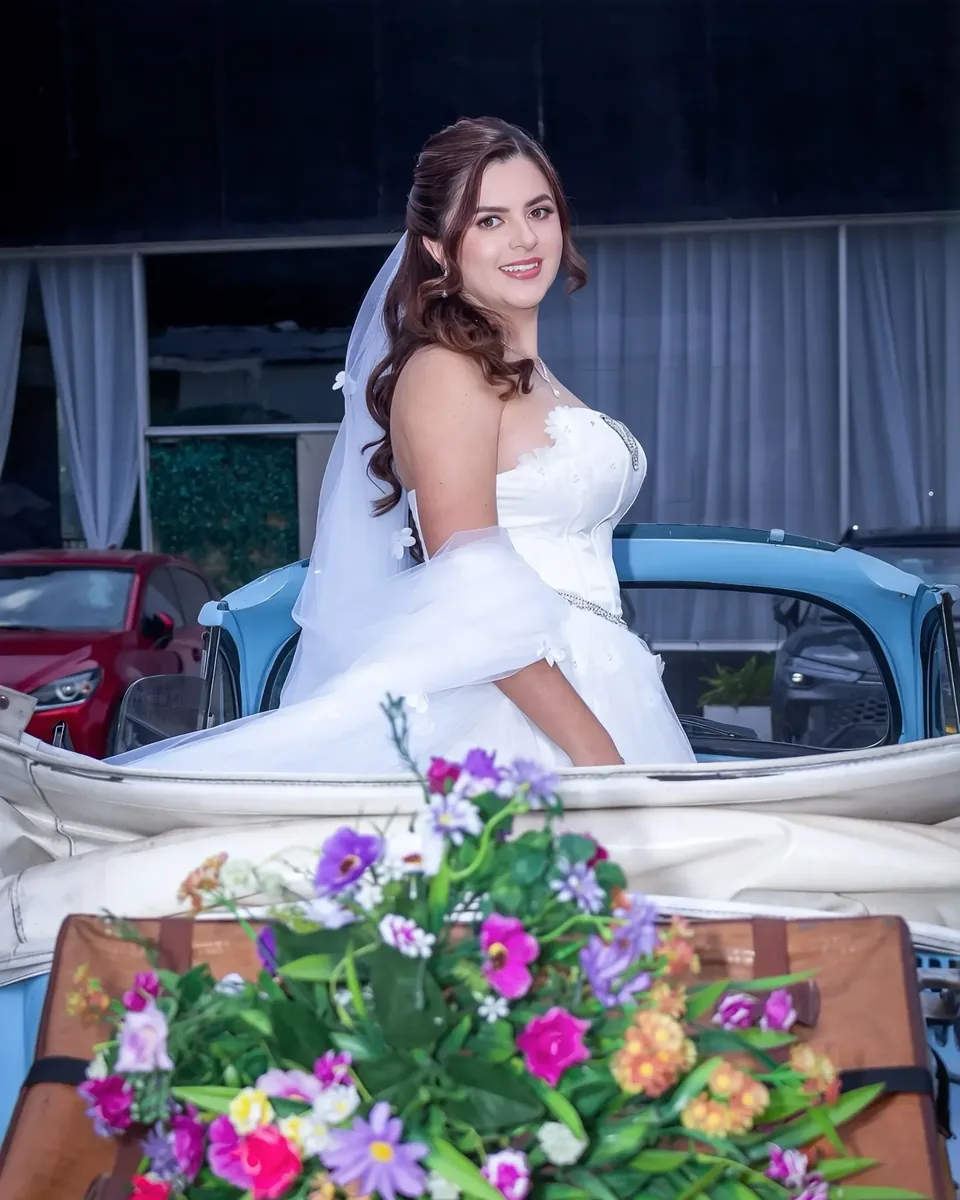 Kelly con maquillaje de novia luminoso, semirrecogido en ondas y velo, de pie en un auto clásico azul
