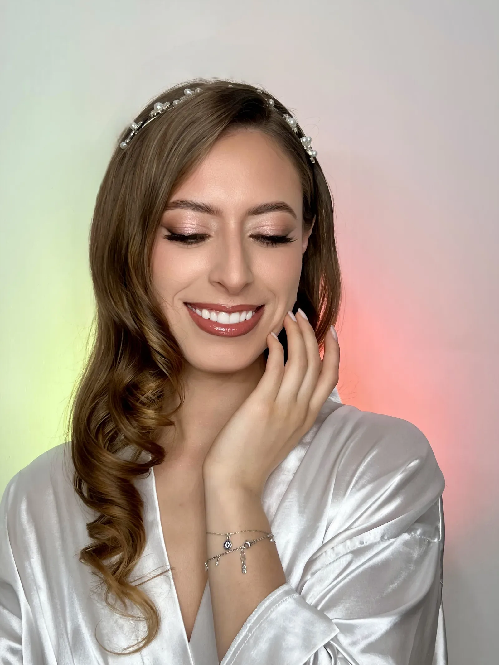 Lau sonríe con ojos cerrados y maquillaje de novia cálido, tiara de perlas y bata de satín blanca