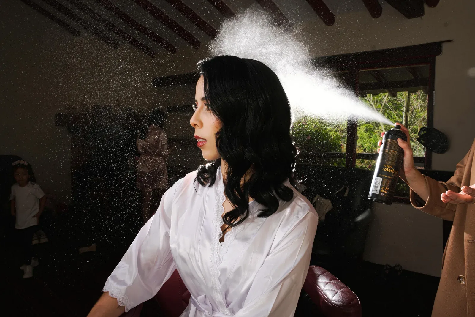 Laura de perfil con maquillaje de novia y labios rojos mientras le aplican fijador sobre sus ondas negras