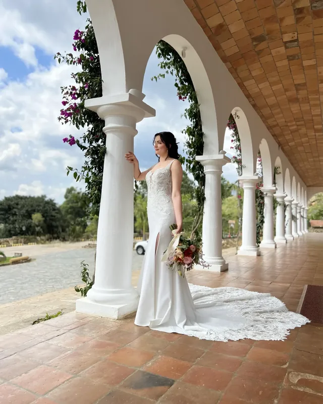 Bride in a white gown holding a flower bouquet beside an arched colonnade.