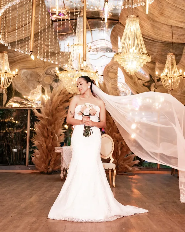 Bride in a lace gown holding a bouquet as her veil billows beneath chandeliers.