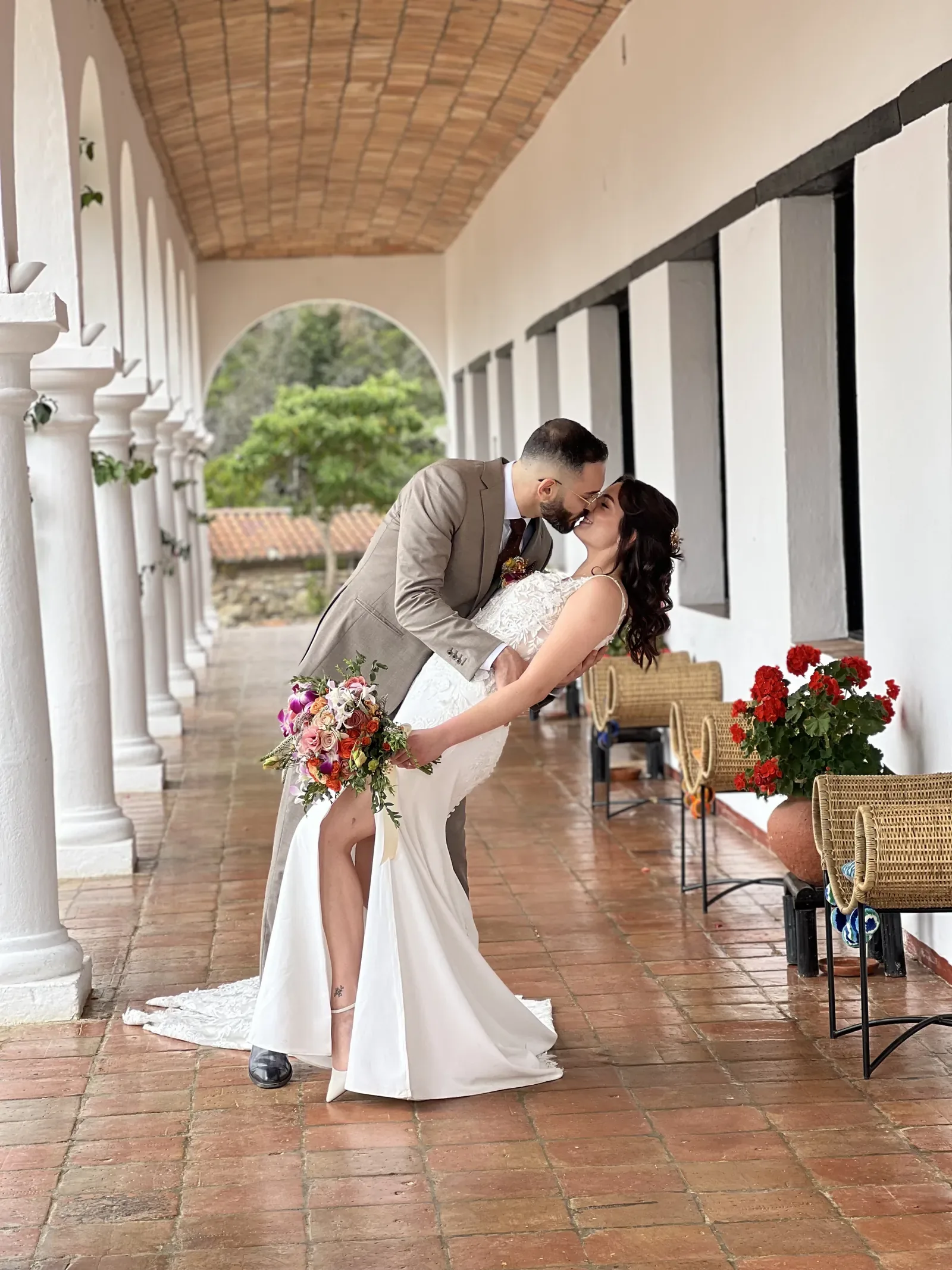 Laura con maquillaje de novia y ondas largas besa al novio en un corredor de arcos con ramo colorido