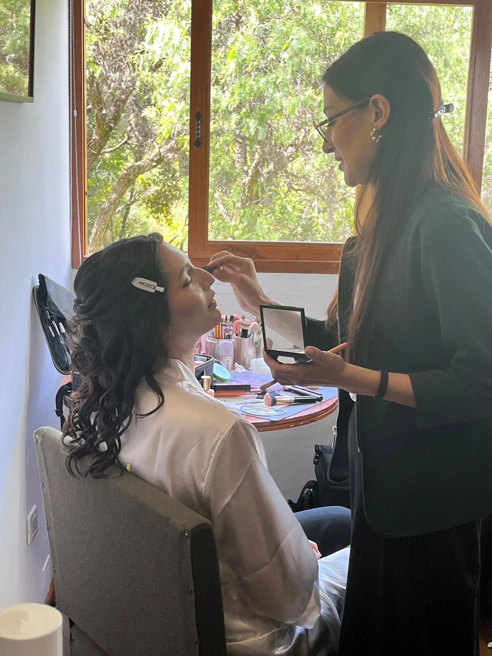 Laura en bata blanca con ondas largas recibe su maquillaje de novia junto a una ventana