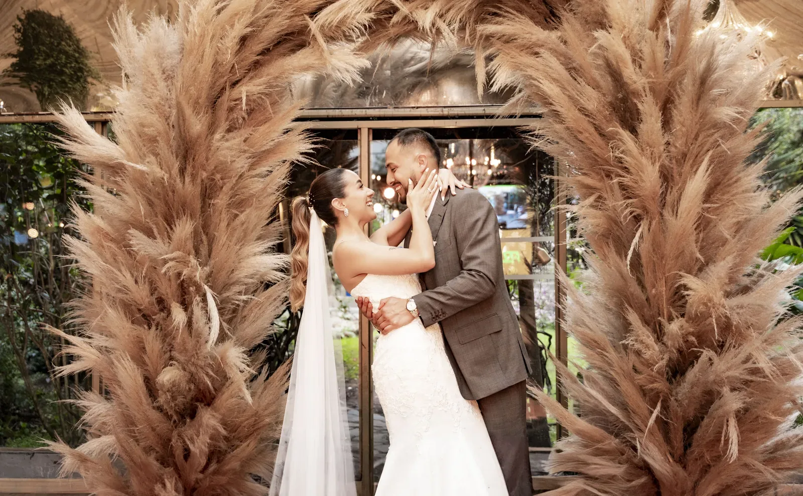 Laura ríe frente al novio bajo un arco de pampa, con maquillaje de novia, cola alta con ondas y velo
