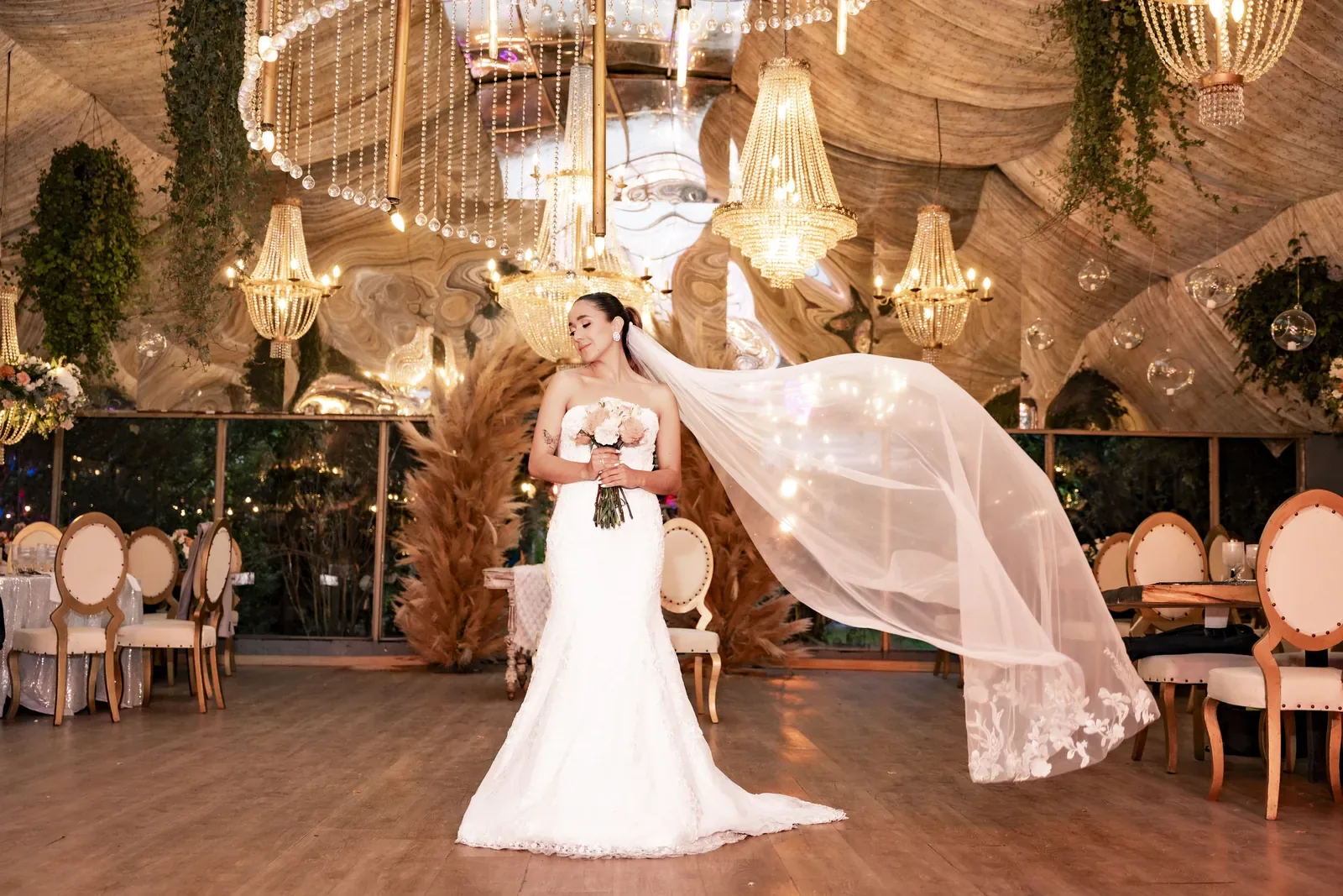 Laura con maquillaje de novia, ramo de rosas y velo largo al viento en un salón con candelabros de cristal