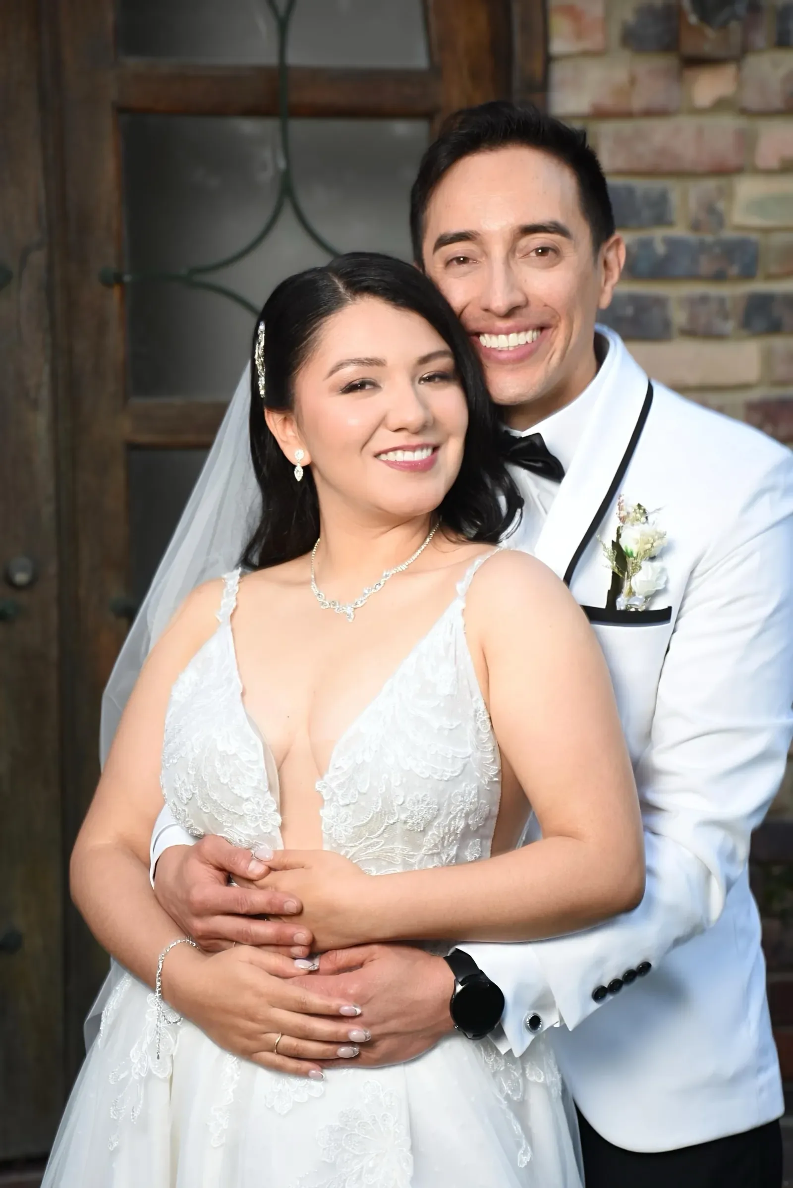 Luisa sonríe abrazada por el novio de saco blanco, con maquillaje de novia luminoso, velo y collar de cristales