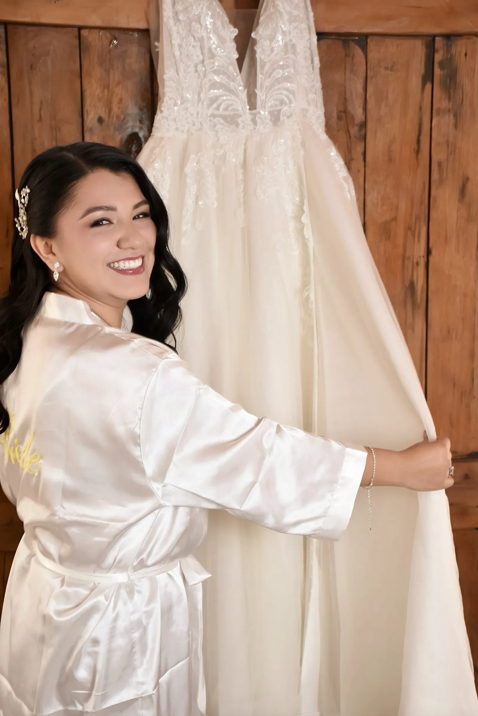 Luisa en bata de satín sonríe junto a su vestido colgado, ya con maquillaje de novia y ondas sueltas