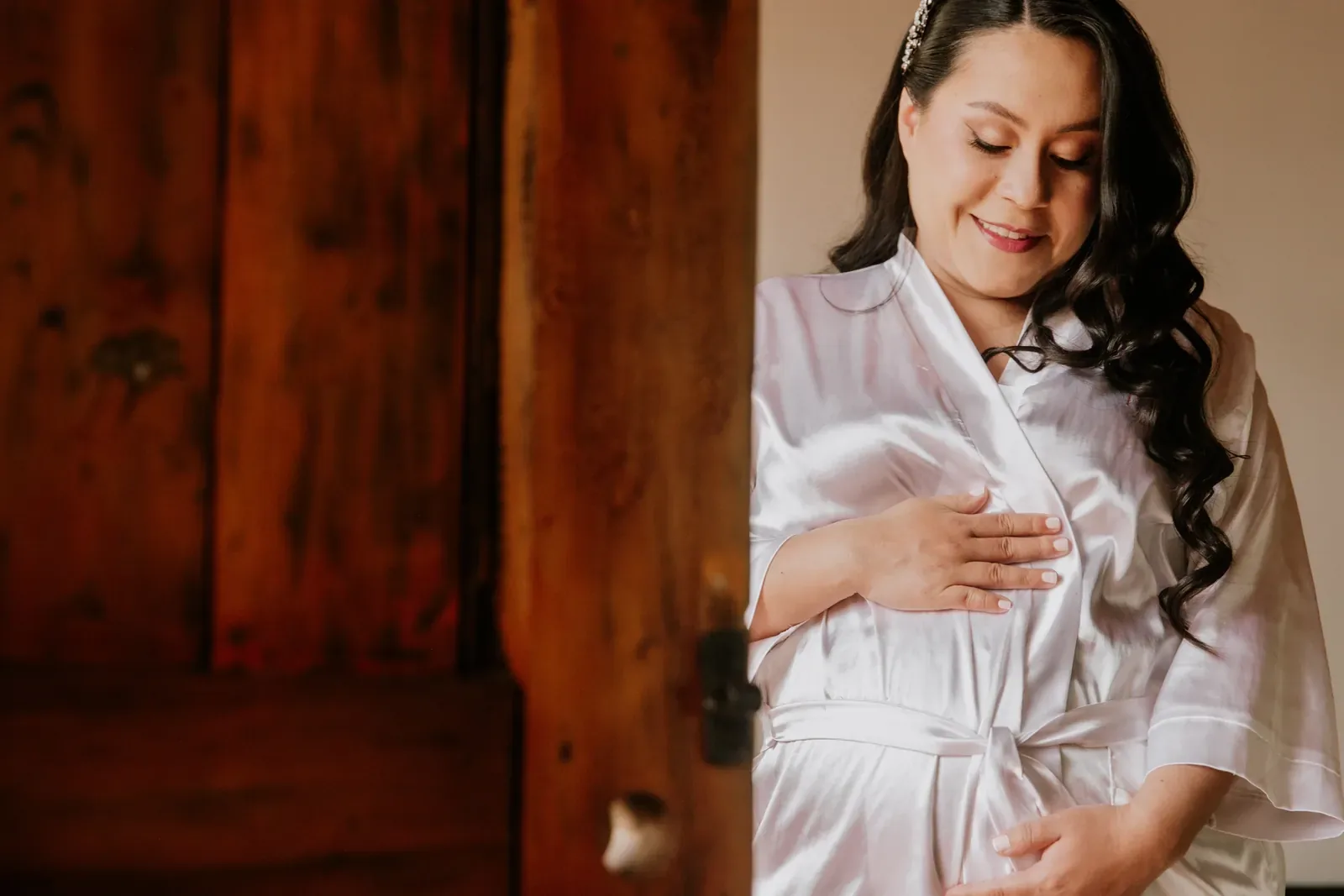 Luisa en bata de satín, con ondas largas y maquillaje de novia de labios rosados, junto a una puerta de madera