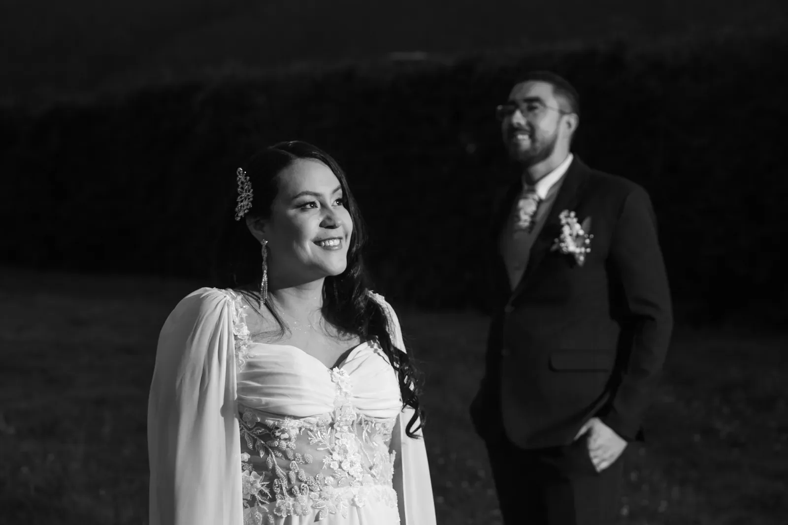 Luisa sonríe con maquillaje de novia, tocado de cristales y vestido con capa de gasa; el novio al fondo