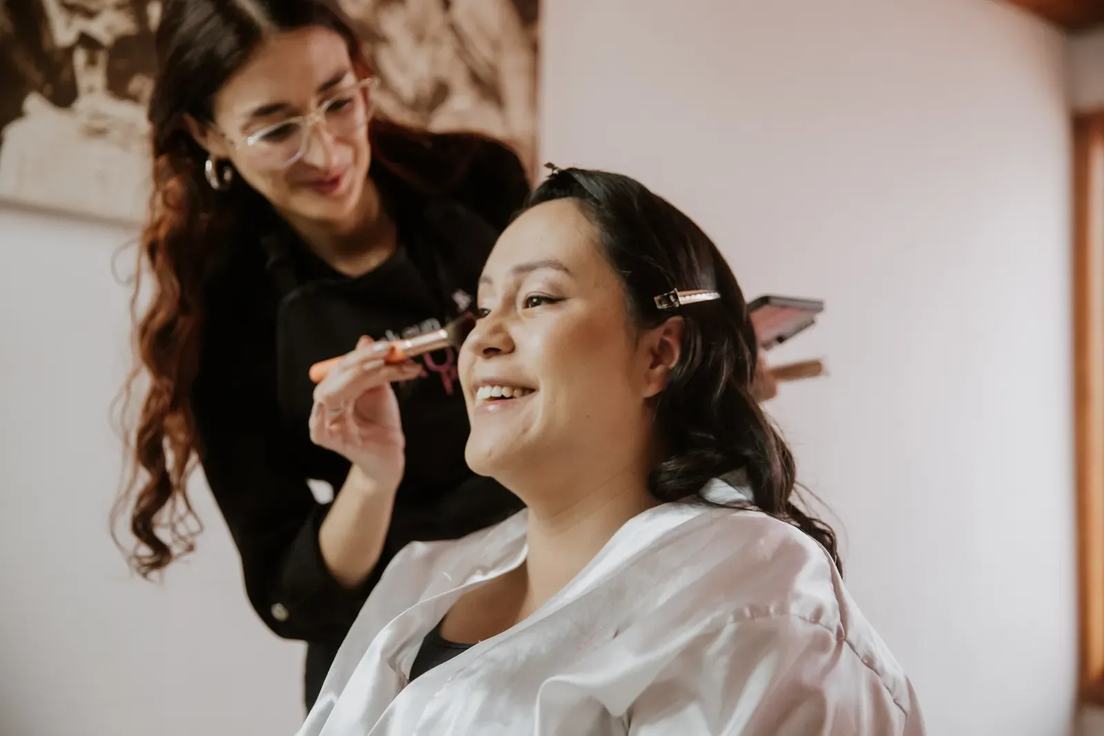 Luisa ríe en bata blanca mientras una maquilladora le aplica rubor con brocha en su maquillaje de novia