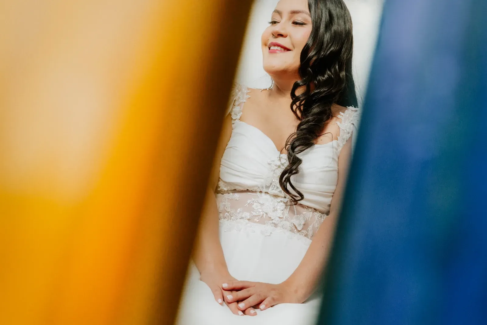 Luisa sonríe con maquillaje de novia y labios coral, ondas largas y vestido de encaje, entre telas de color