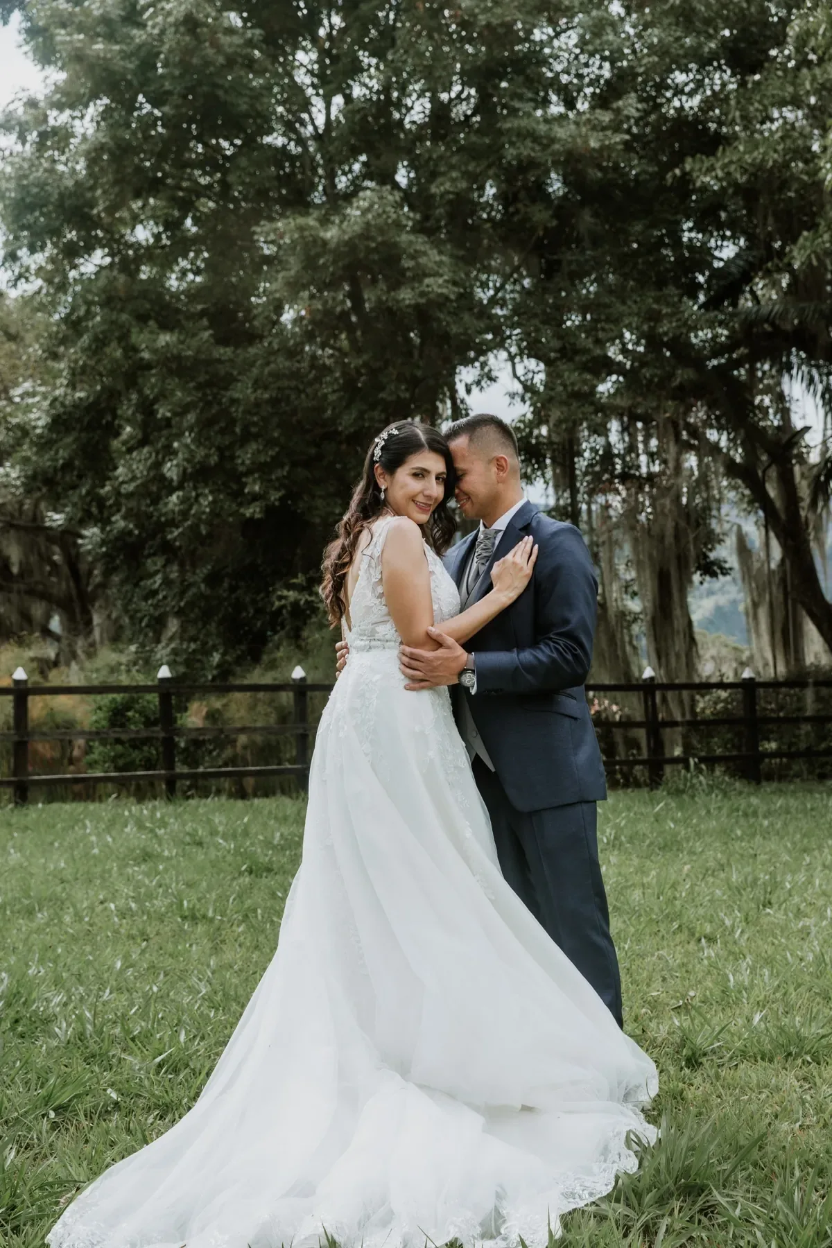 Luisa con maquillaje de novia, ondas y tocado brillante abraza al novio en un prado con árboles