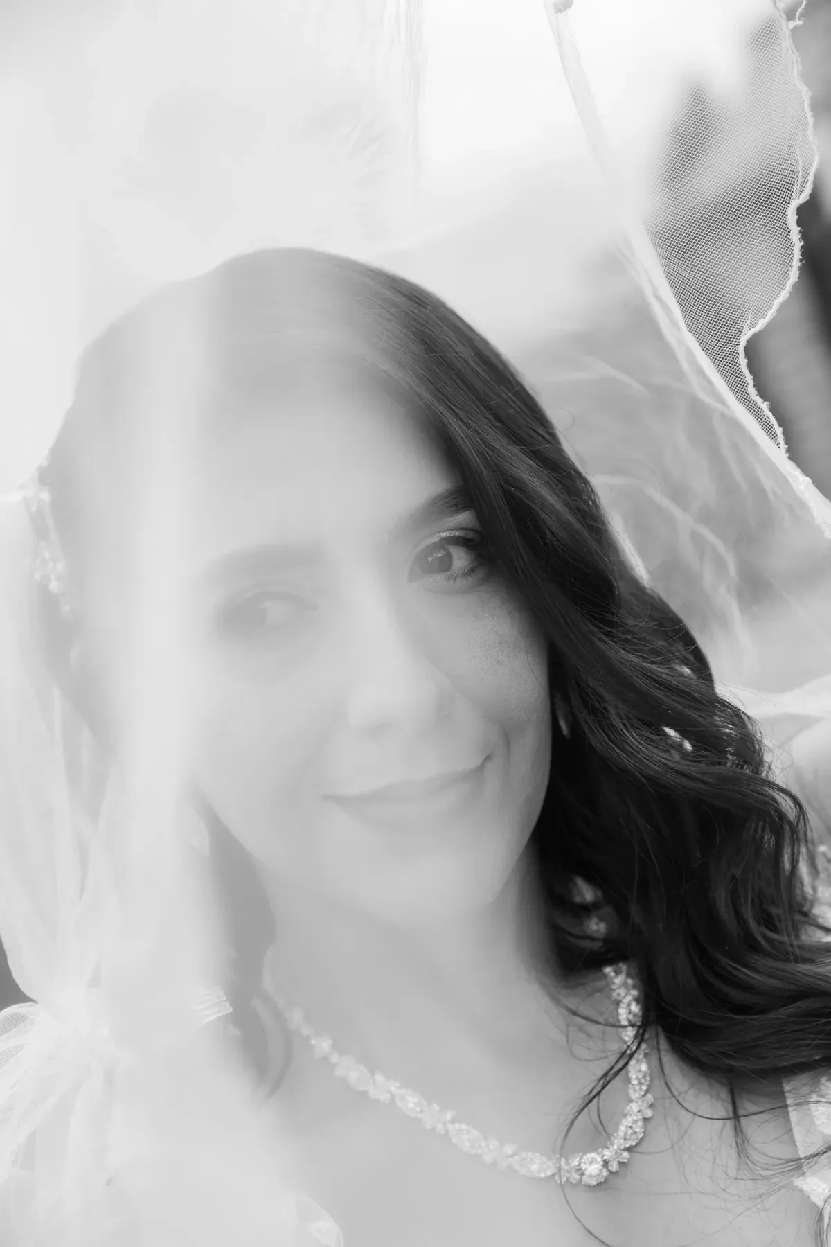 Luisa sonríe bajo el velo con maquillaje de novia suave, ondas sueltas y collar de cristales