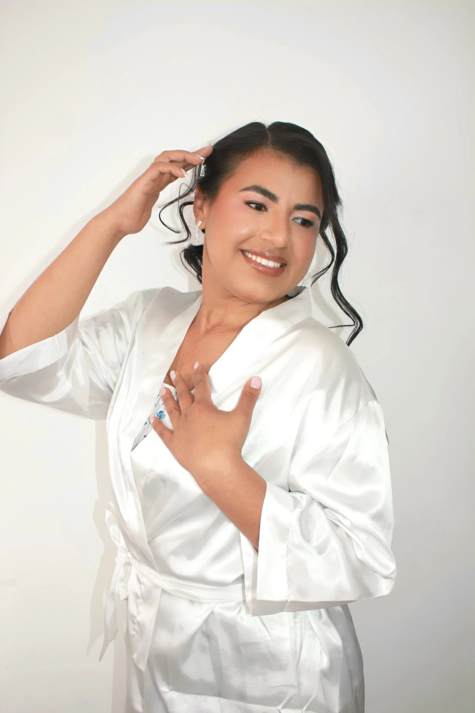 Novia sonriente con maquillaje rosado y bata blanca de satén, acomodándose el cabello.