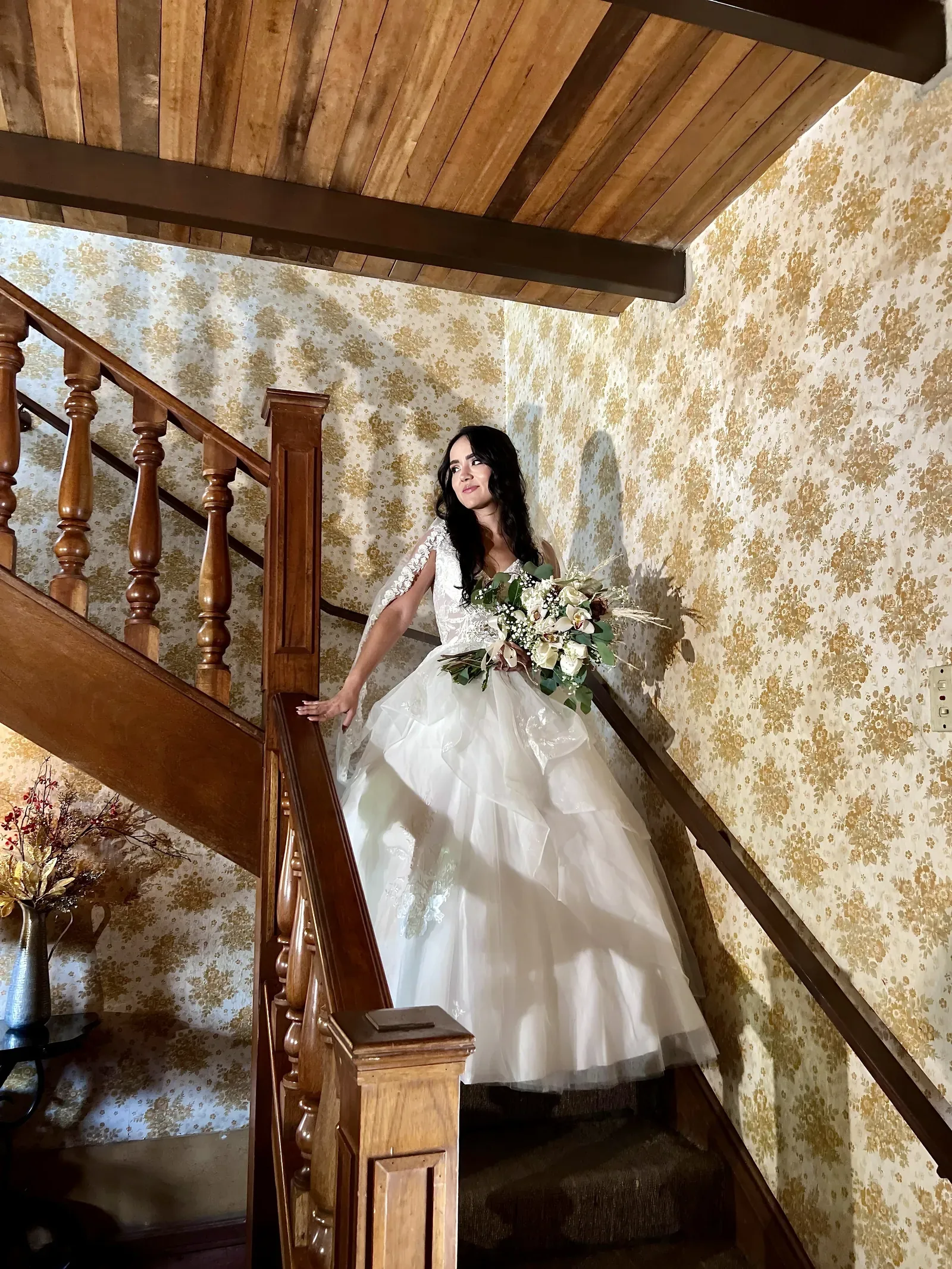 Natalie con maquillaje de novia bajando una escalera de madera con ramo de flores blancas y vestido de tul