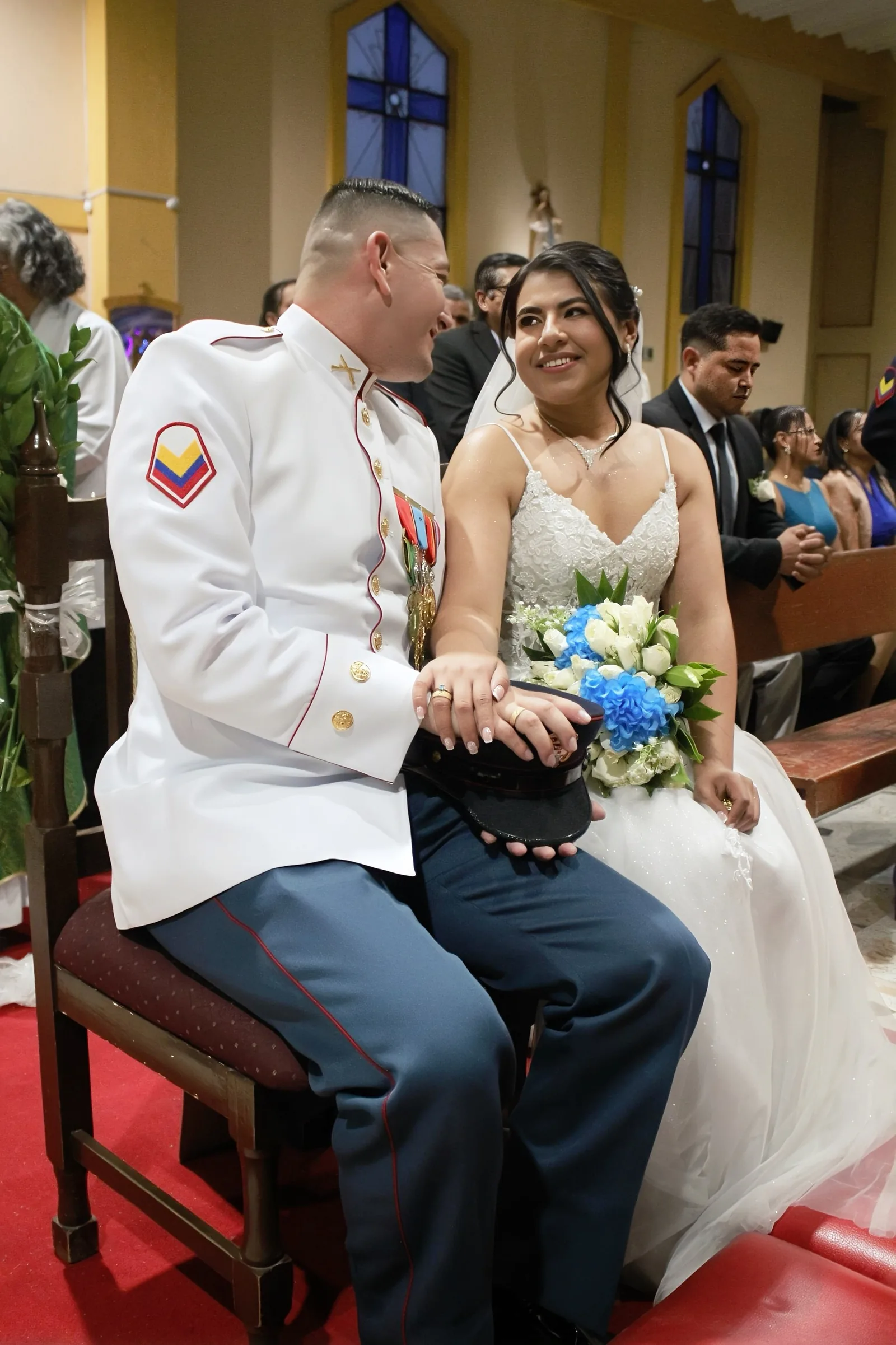 Novia con ramo de flores azules y blancas sentada junto a su pareja en una iglesia.
