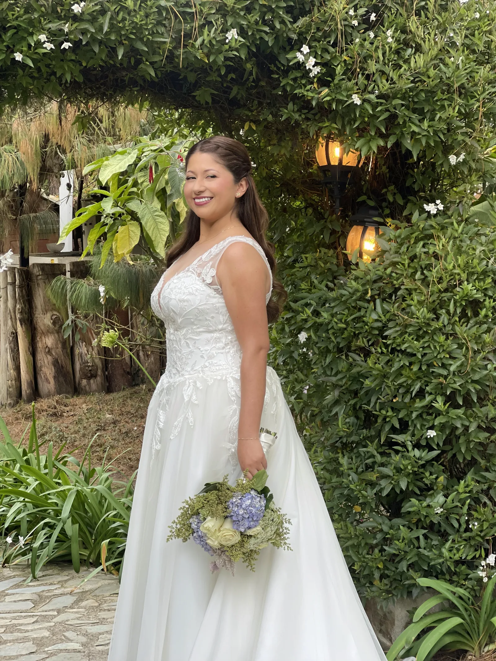 Smiling bride in a white lace gown holding a bouquet of hydrangeas in a leafy garden.
