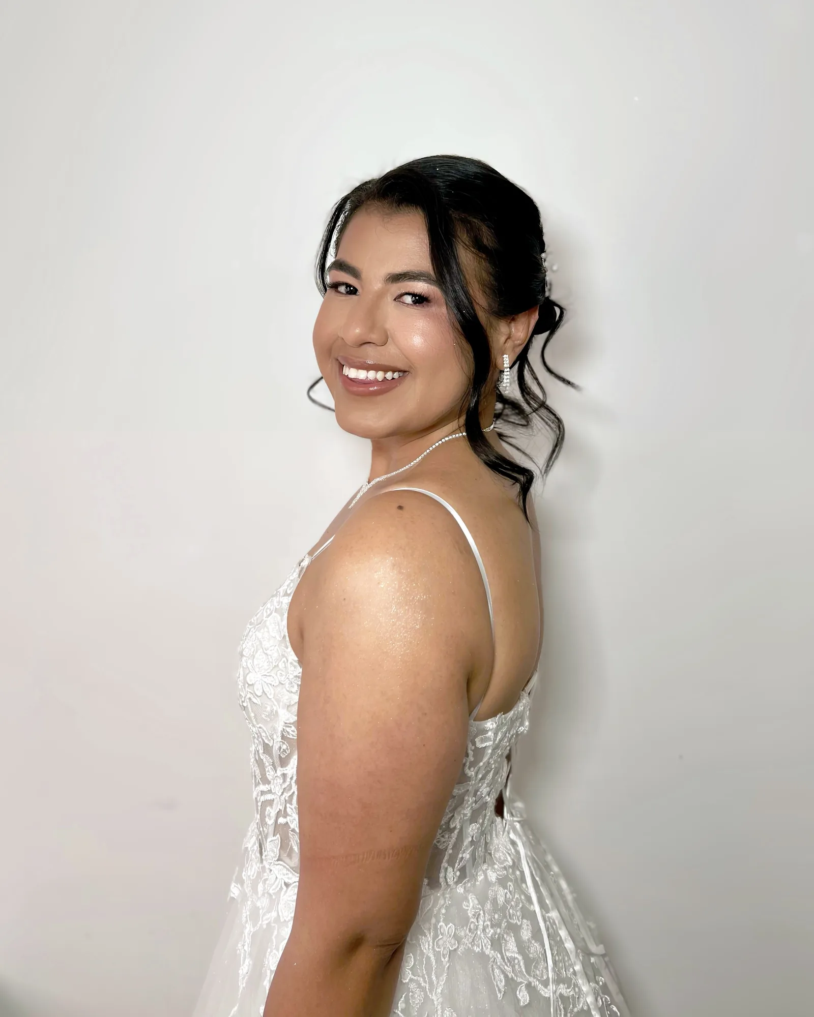 Novia sonriente con maquillaje luminoso, peinado recogido y vestido de encaje.