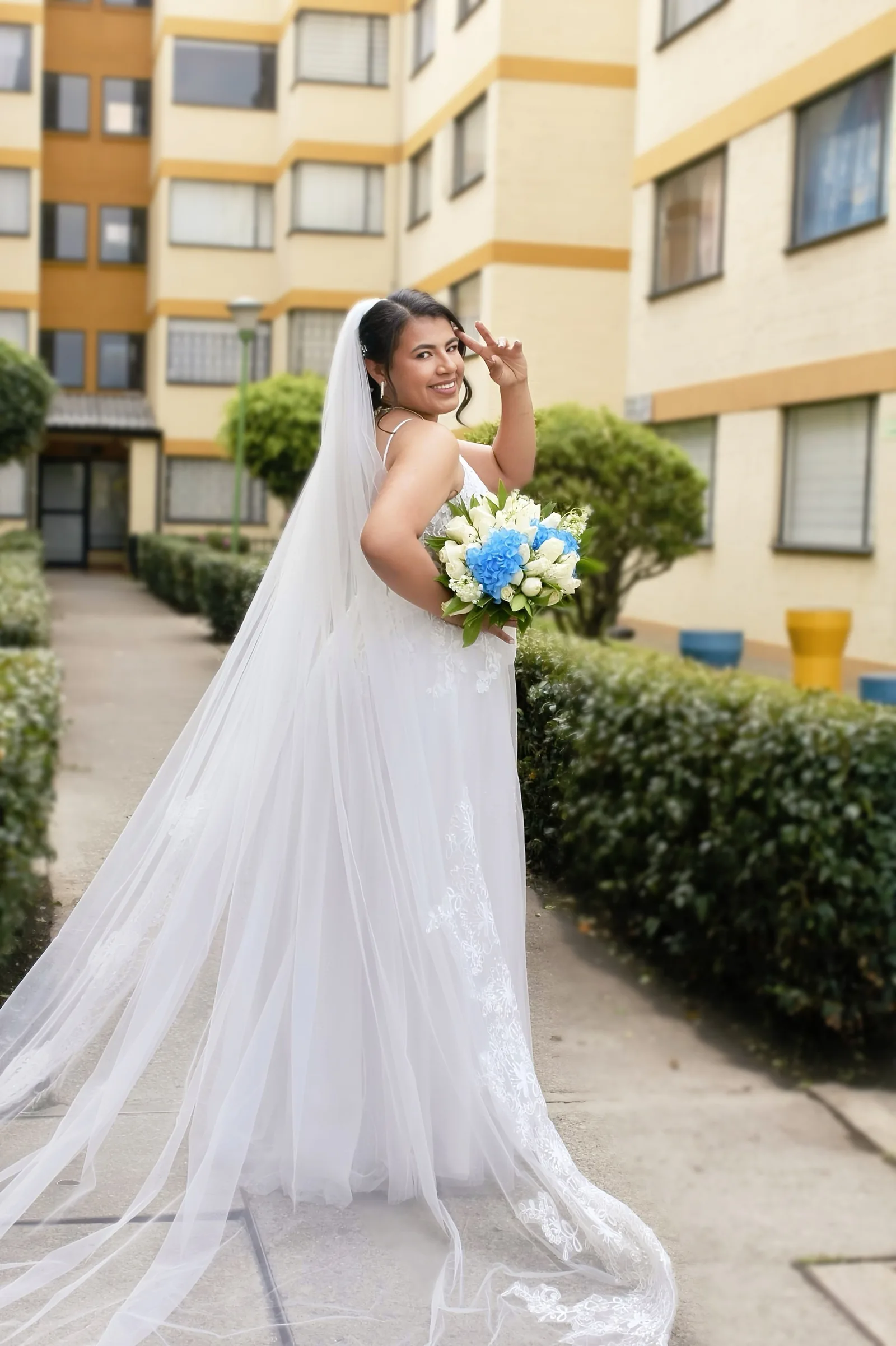 Novia sonriente con vestido de encaje y velo largo, sosteniendo un ramo de flores blancas y azules.