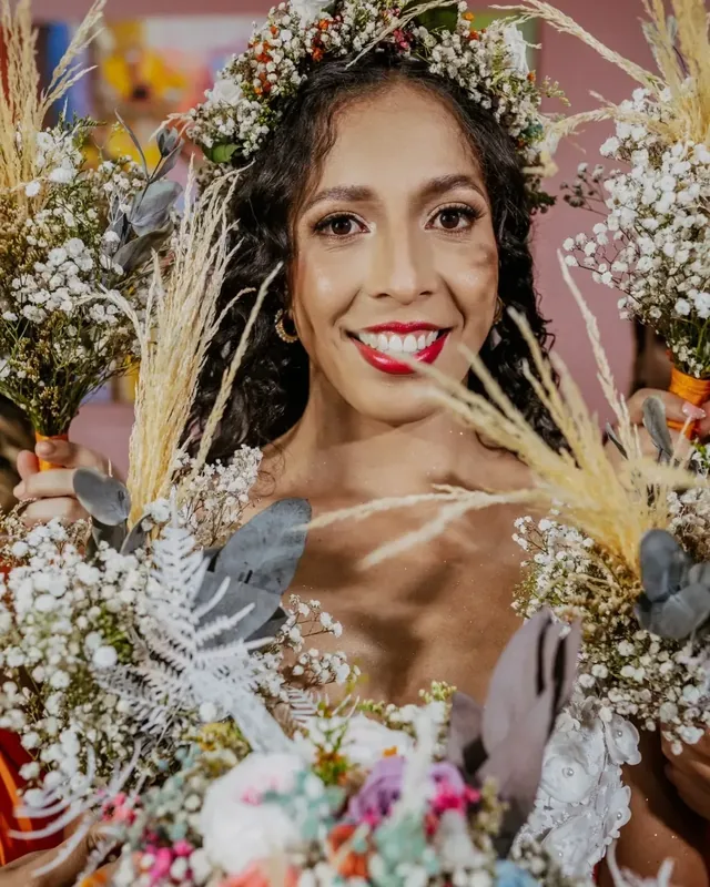 Stefa sonríe con maquillaje de novia de labios rojos y corona de flores, rodeada de ramos de paniculata