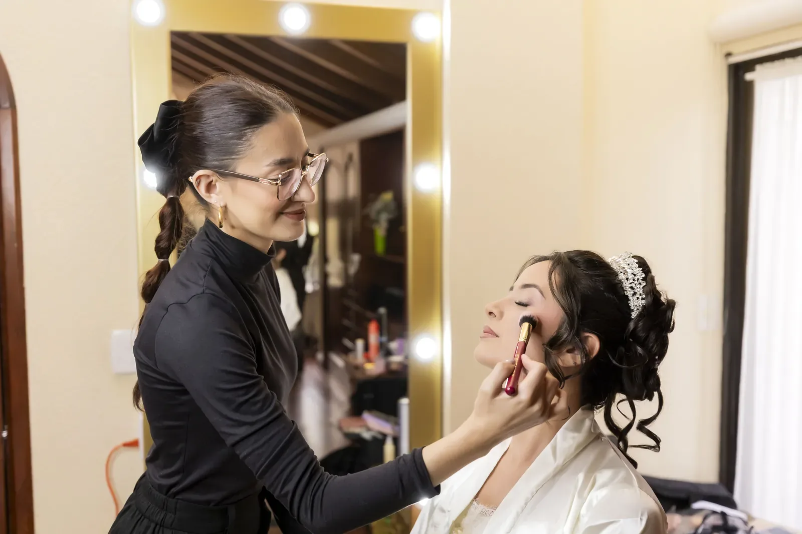 Tatiana con tiara y recogido en rizos recibe su maquillaje de novia con brocha frente a un espejo
