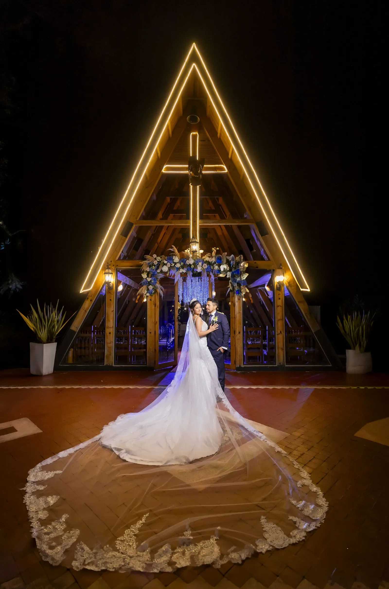 Tatiana con maquillaje de novia, tiara y velo catedral junto al novio frente a una capilla iluminada