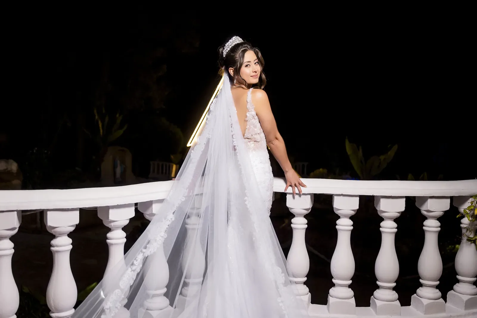 Tatiana mira sobre el hombro con maquillaje de novia, tiara y velo largo en una balaustrada de noche