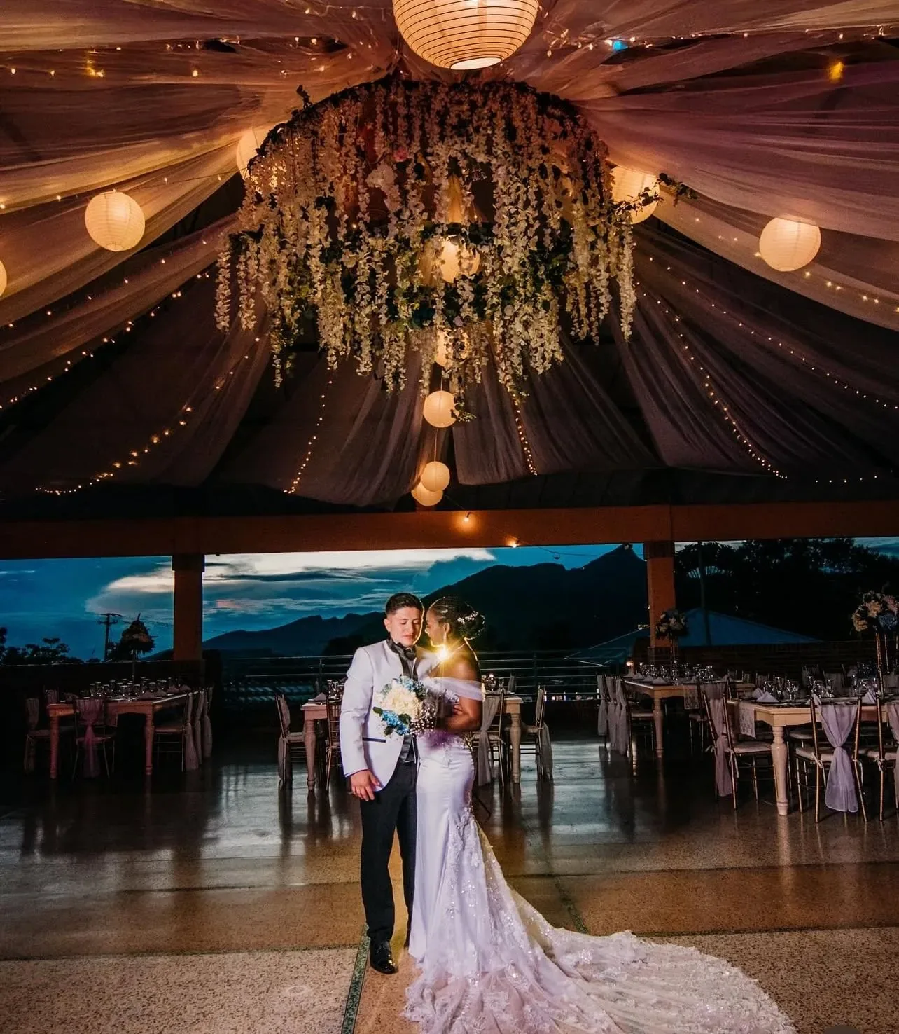 Trudy con maquillaje de novia junto a su pareja bajo faroles y flores colgantes al anochecer