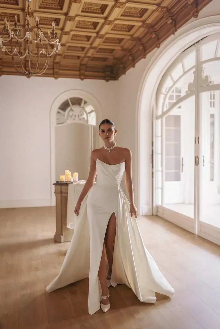 Modelo con vestido blanco de novia con abertura, en un salón con ventanales.