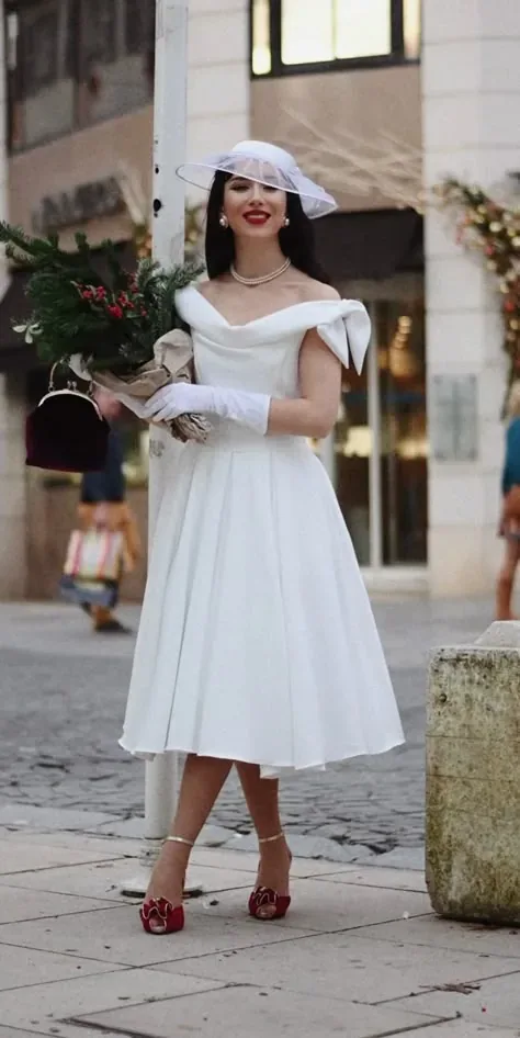 Novia con vestido blanco de largo midi, velo tipo sombrero y labios rojos, sosteniendo un ramo.